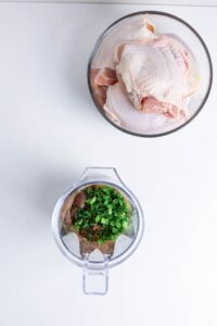 A glass bowl of raw chicken pieces and a blender containing sauce ingredients and chopped green onions on a white surface.