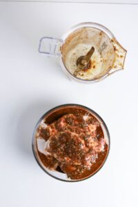 A food processor with residue sits beside a bowl of raw meat covered in a red marinade on a white surface.