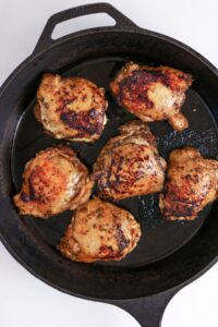 Six seasoned and seared chicken thighs cooking in a black cast iron skillet on a white surface.