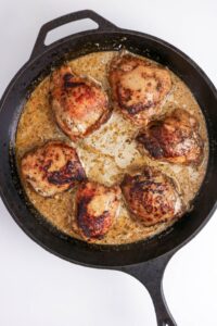 Six browned chicken thighs cooking in a creamy sauce inside a black cast iron skillet on a white background.