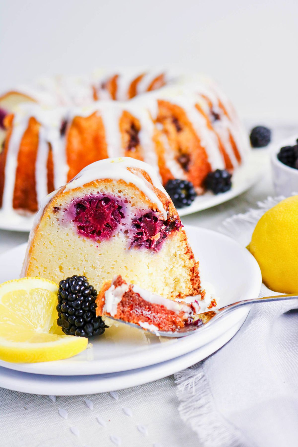 A slice of glazed berry bundt cake on a white plate with lemon wedges and blackberries, with the whole cake in the background.
