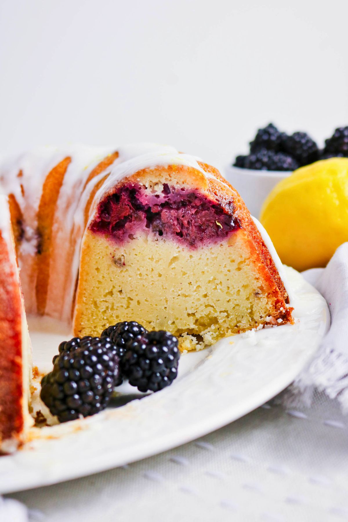 A sliced bundt cake with a layer of blackberries inside, topped with white icing, served with fresh blackberries and a lemon in the background.