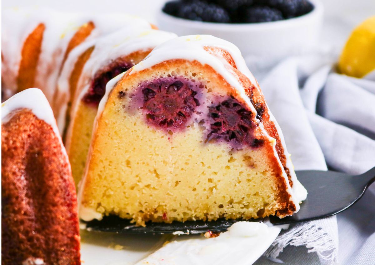 A slice of bundt cake with blackberries inside, topped with white icing, is being served with a cake spatula. A bowl of blackberries and a lemon are in the background.