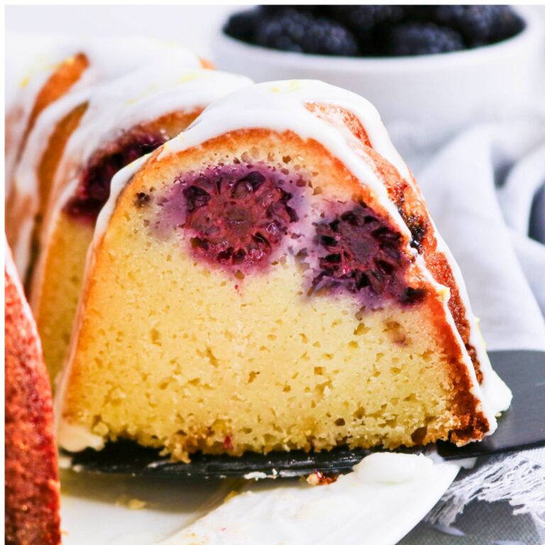 A close-up of a sliced bundt cake with blackberries baked inside and white icing drizzled on top, with a bowl of blackberries in the background.
