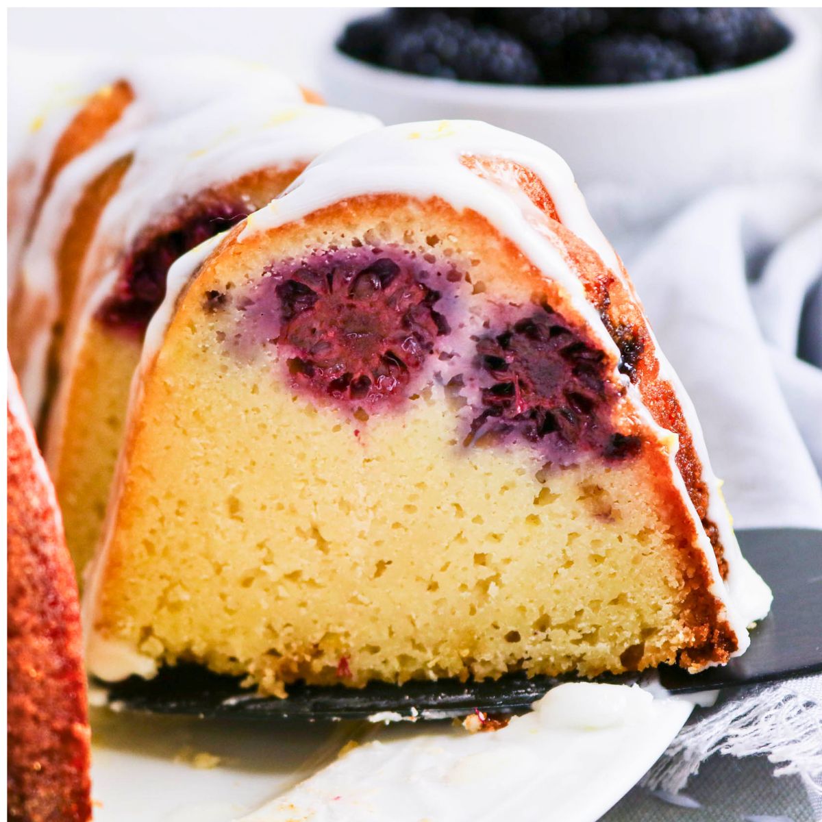 A close-up of a sliced bundt cake with blackberries baked inside and white icing drizzled on top, with a bowl of blackberries in the background.