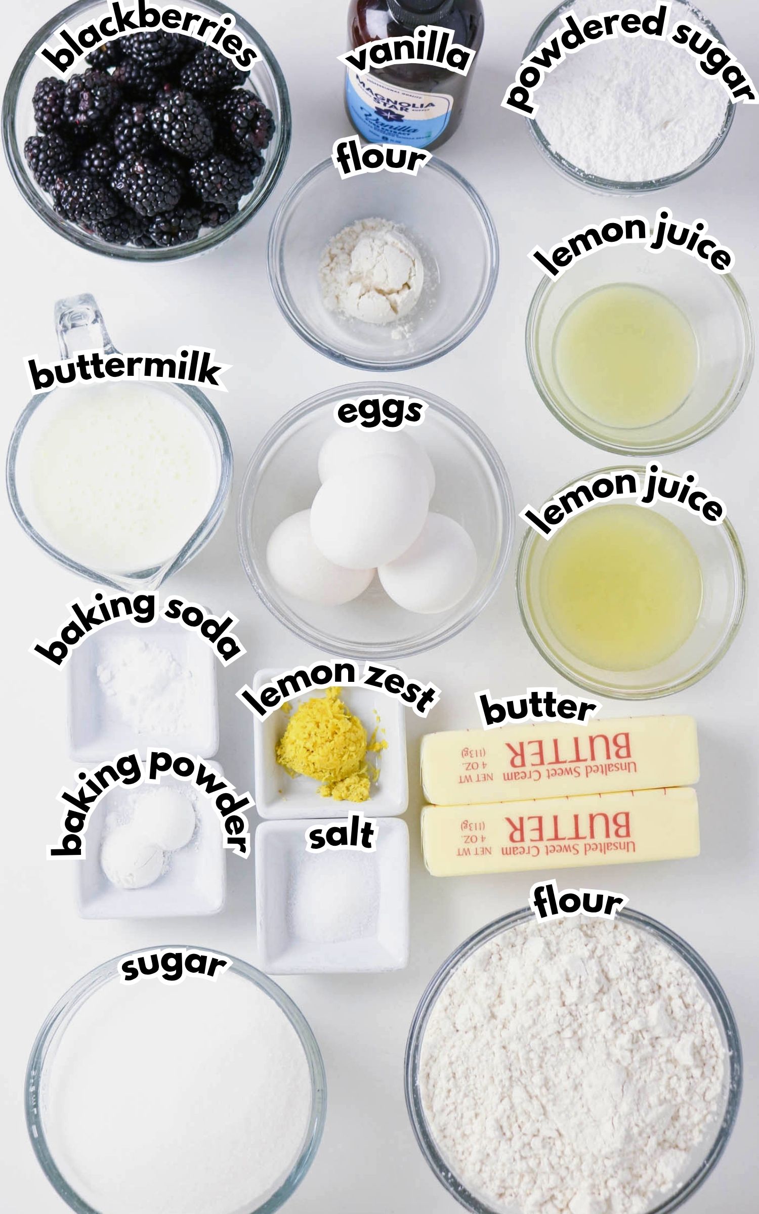 A top-down view of labeled baking ingredients in bowls and containers, including blackberries, eggs, butter, flour, sugar, buttermilk, baking powder, baking soda, salt, vanilla, lemon zest, and lemon juice.