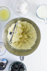 A mixing bowl with creamed butter and sugar, surrounded by eggs, milk, blackberries, flour, vanilla extract, and a bowl of egg whites on a white surface.