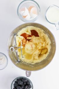 A metal mixing bowl with creamed ingredients, eggs, zest, and liquid, surrounded by bowls of eggshells, flour, blackberries, and a measuring cup of milk.