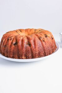 A golden-brown bundt cake with visible raisins sits on a white plate against a plain white background.