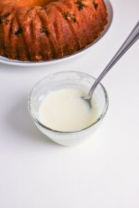 A glass bowl of white glaze with a spoon inside sits in front of a partially visible bundt cake on a white surface.