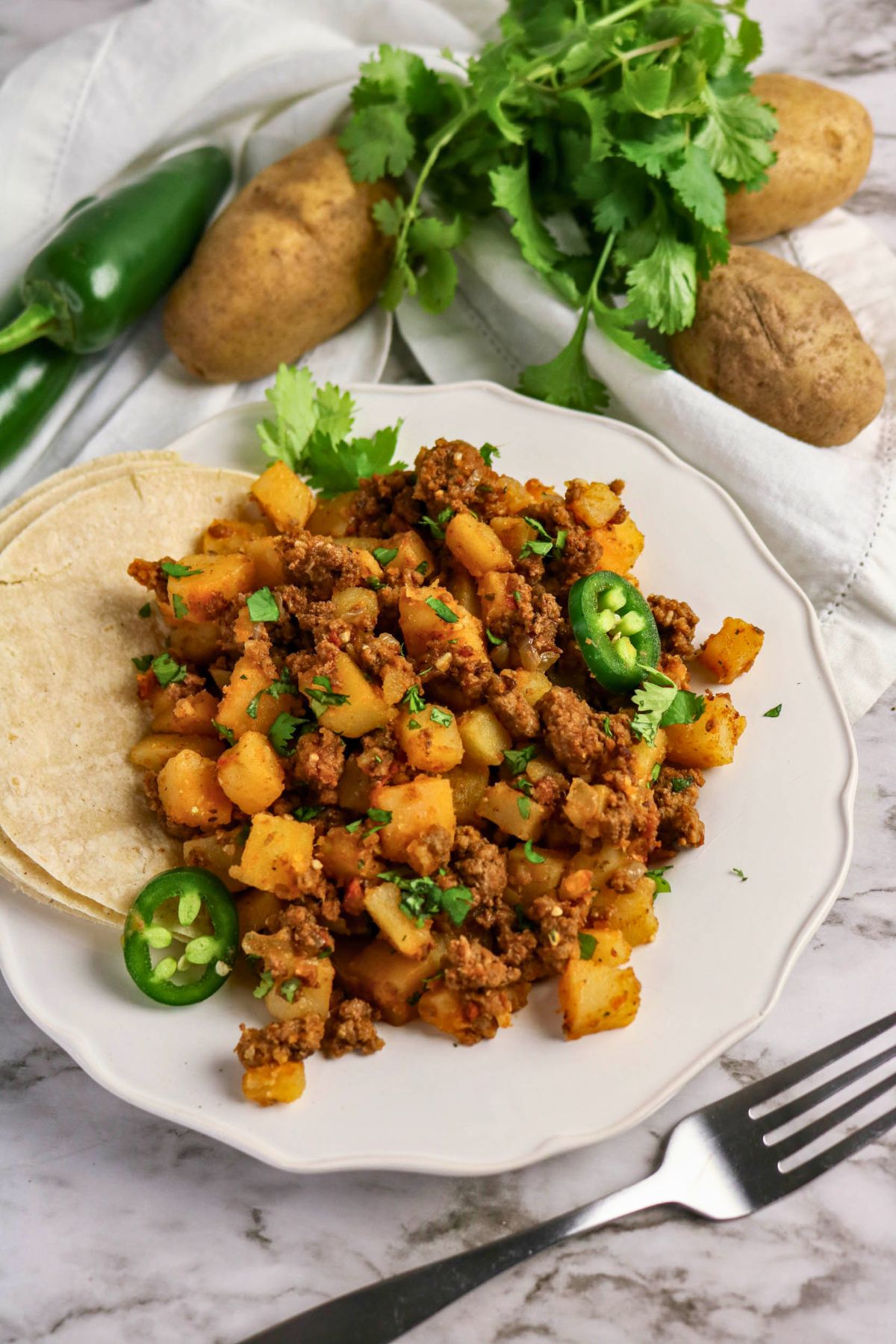 A plate of ground beef and potato hash garnished with cilantro and sliced jalapeños, served with corn tortillas. Fresh potatoes, cilantro, and a jalapeño are in the background.