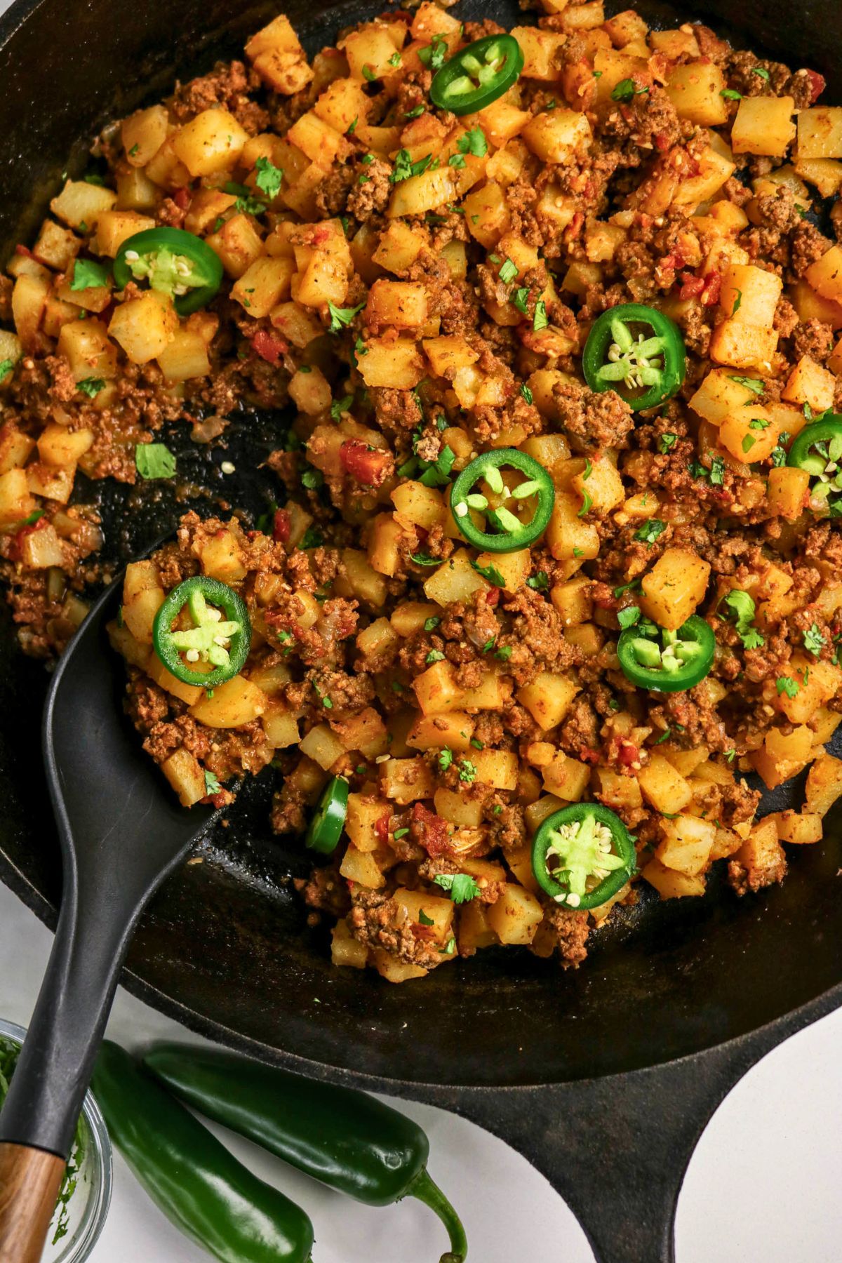 A skillet filled with cooked diced potatoes, ground meat, tomatoes, and sliced jalapeños, with a black serving spoon and two whole jalapeños beside the pan.