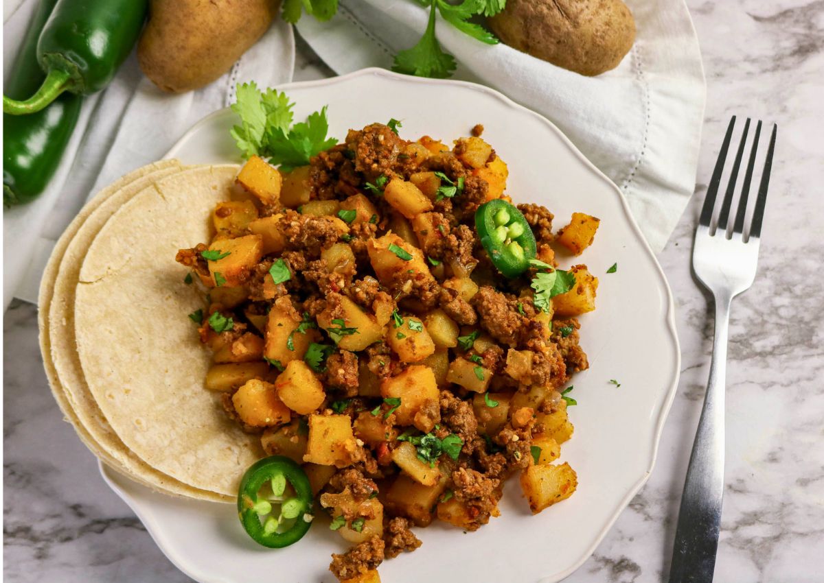 A plate of ground beef and potato hash garnished with sliced jalapeños and cilantro, served with corn tortillas; potatoes and jalapeños in the background.
