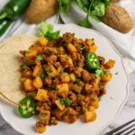 A plate of ground beef and diced potatoes garnished with cilantro and jalapeño slices, served with corn tortillas. Potatoes, jalapeños, and cilantro are in the background.