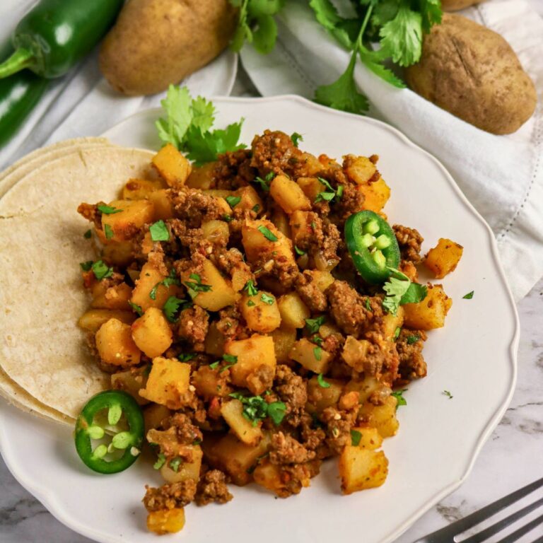 A plate of ground beef and diced potatoes garnished with cilantro and jalapeño slices, served with corn tortillas. Potatoes, jalapeños, and cilantro are in the background.