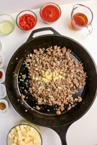 A cast iron skillet with cooked ground beef and chopped onions in the center, surrounded by bowls of diced potatoes, sauces, seasonings, and liquids on a white surface.