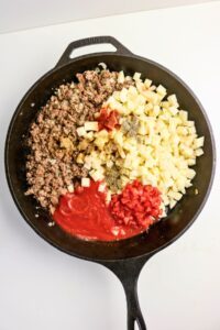 A cast iron skillet with ground beef, diced potatoes, tomato sauce, diced tomatoes, and assorted seasonings, all unmixed.