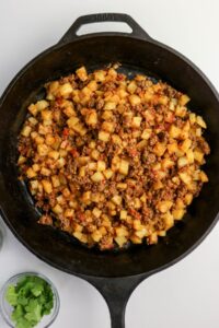 A cast iron skillet filled with cooked ground meat, diced potatoes, and seasonings sits on a white surface. Small bowls with ingredients are next to the skillet.