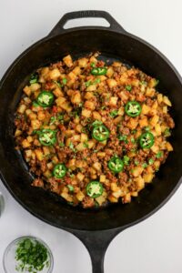 A cast iron skillet filled with diced potatoes and ground meat, garnished with sliced jalapeños and chopped herbs.