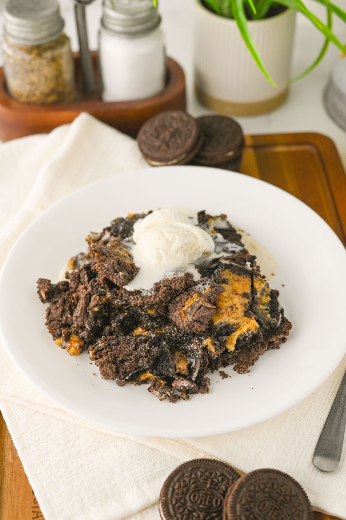 A plate of chocolate Oreo dessert topped with a scoop of vanilla ice cream, with whole Oreo cookies and condiments in the background.