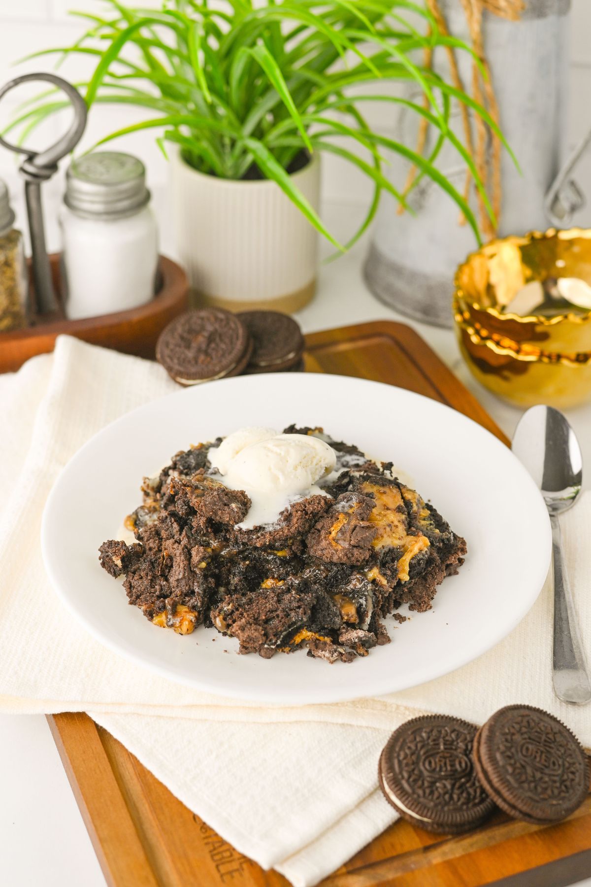 A plate with a serving of Oreo dessert topped with vanilla ice cream sits on a white cloth, with Oreo cookies and kitchen items in the background.