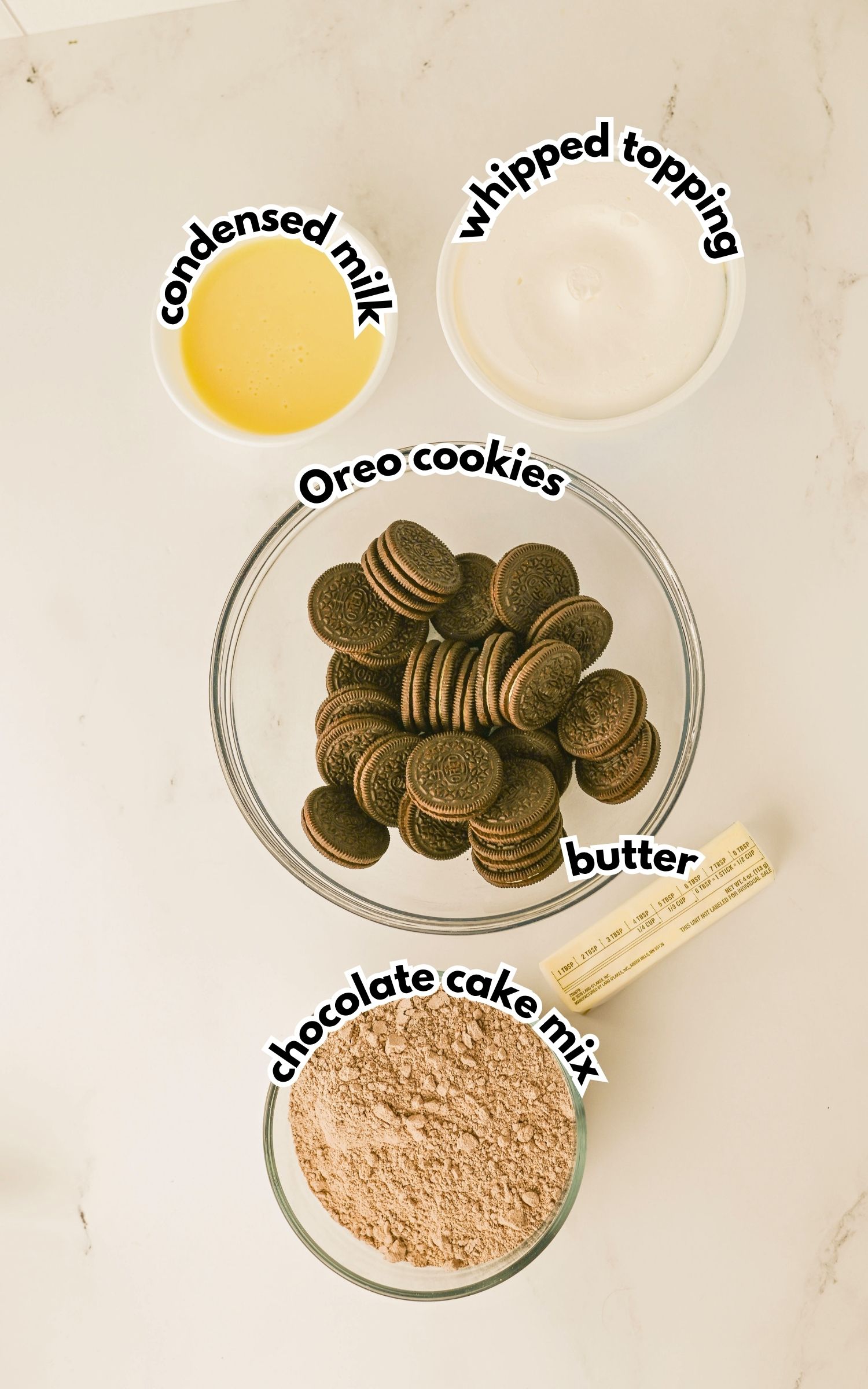 Five labeled ingredients on a countertop: condensed milk, whipped topping, Oreo cookies in a bowl, a stick of butter, and chocolate cake mix in a bowl.