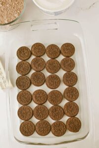 A glass baking dish with a single layer of chocolate sandwich cookies arranged in rows. Baking ingredients are visible in the background.