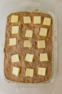 Glass baking dish with a layer of chocolate cake mix topped with evenly spaced squares of butter, ready for baking.