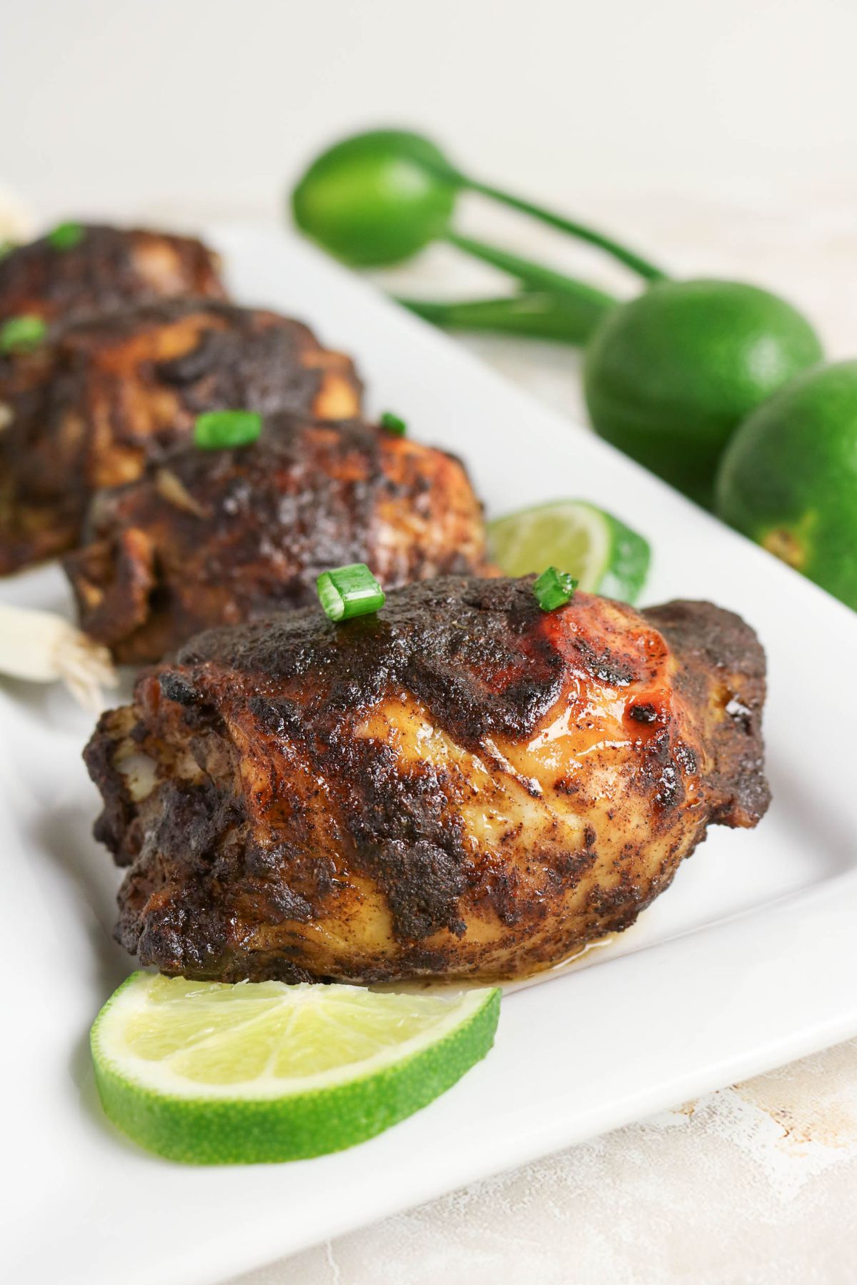 Grilled chicken pieces with a dark spice rub are served on a white plate, garnished with green onions and lime slices, with whole limes in the background.