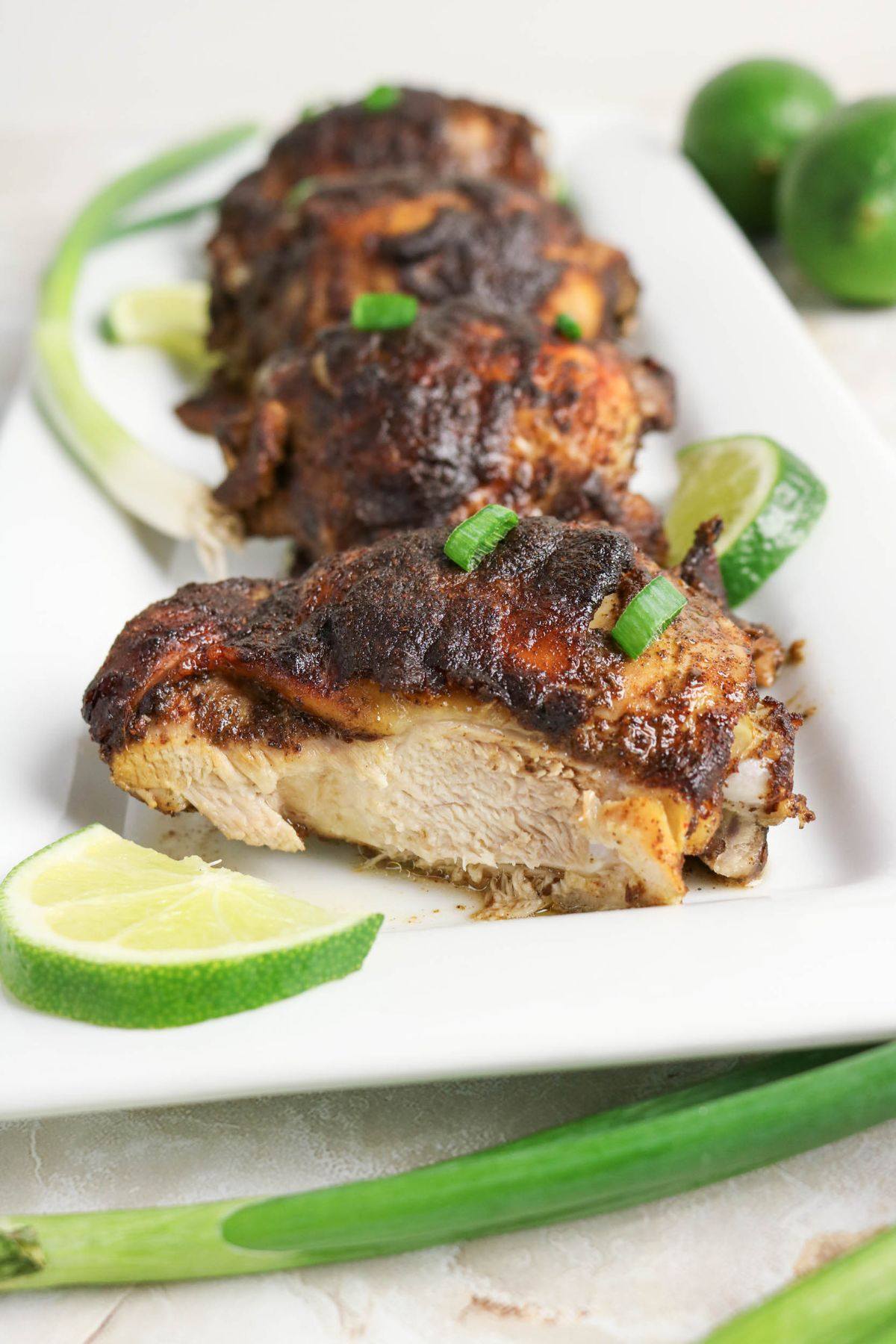 Four pieces of seasoned, cooked chicken on a white plate, garnished with lime wedges and chopped green onions, with whole limes and green onions in the background.