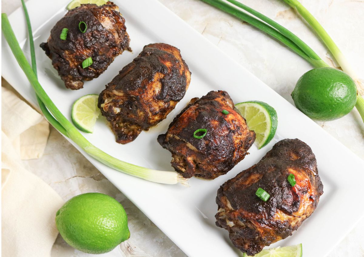 Four pieces of seasoned, cooked chicken are arranged on a white rectangular plate with lime slices, green onions, and whole limes on the side.