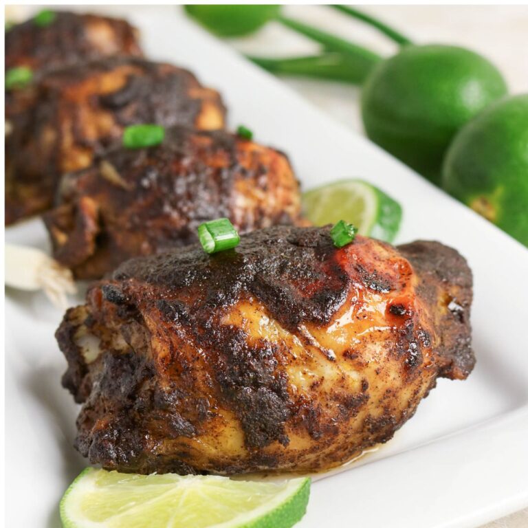 Grilled chicken thighs with a dark, seasoned crust, garnished with chopped green onions and lime slices on a white rectangular plate.