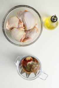 A glass bowl with raw chicken thighs, a glass container of mixed spices, and a bottle of olive oil on a white surface.