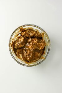 A glass bowl filled with pieces of meat coated in a thick, brown marinade sits on a plain white surface.