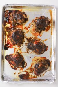 Six cooked chicken thighs on a foil-lined baking sheet, with browned, roasted skin and visible juices and seasonings on the foil.