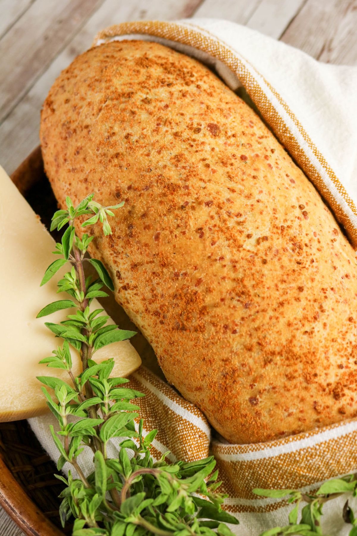 A loaf of bread with a golden-brown crust sits on a cloth, next to fresh green herbs and a slice of cheese.