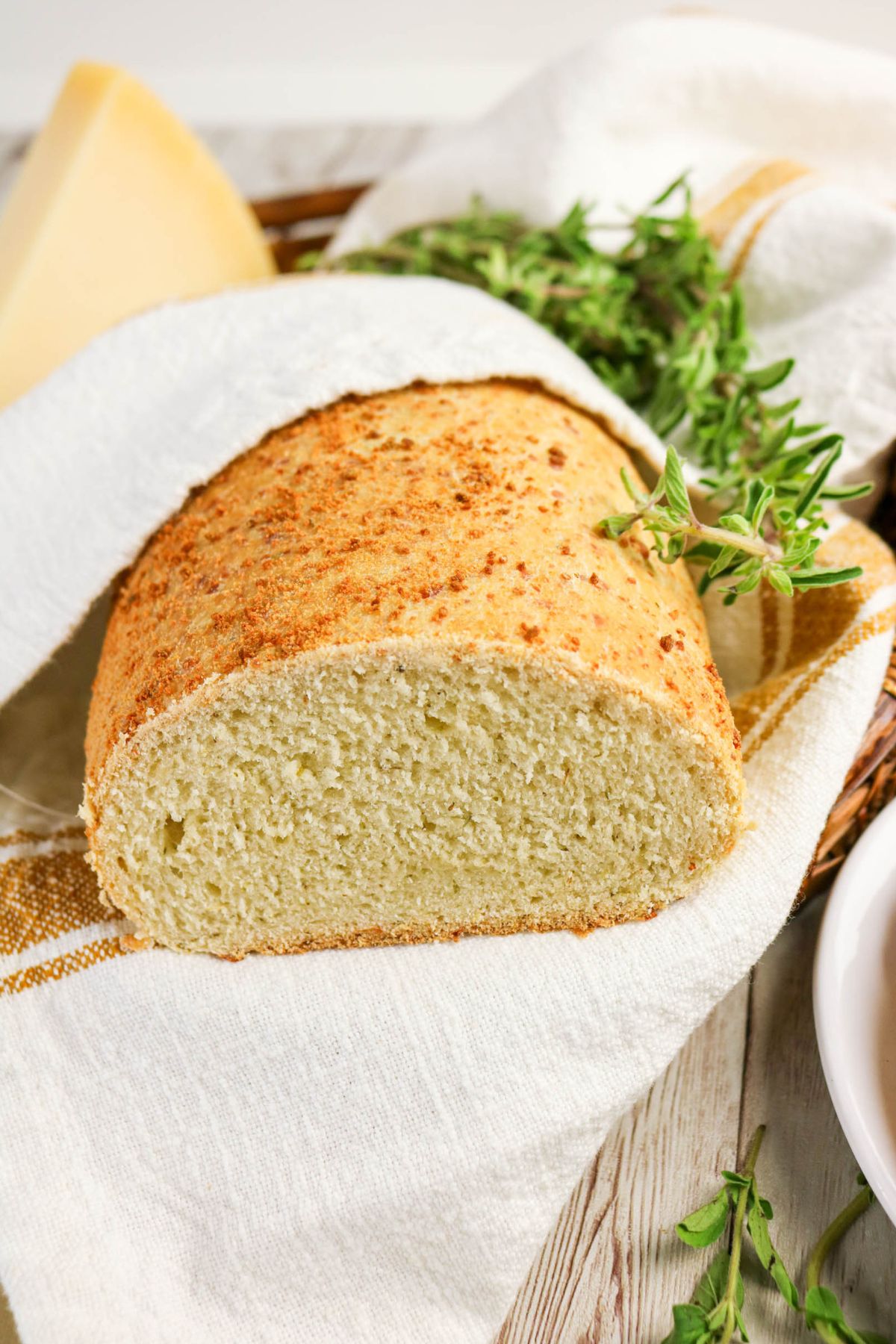 A loaf of bread, partially sliced, rests on a white cloth with sprigs of fresh herbs beside it on a wooden surface.