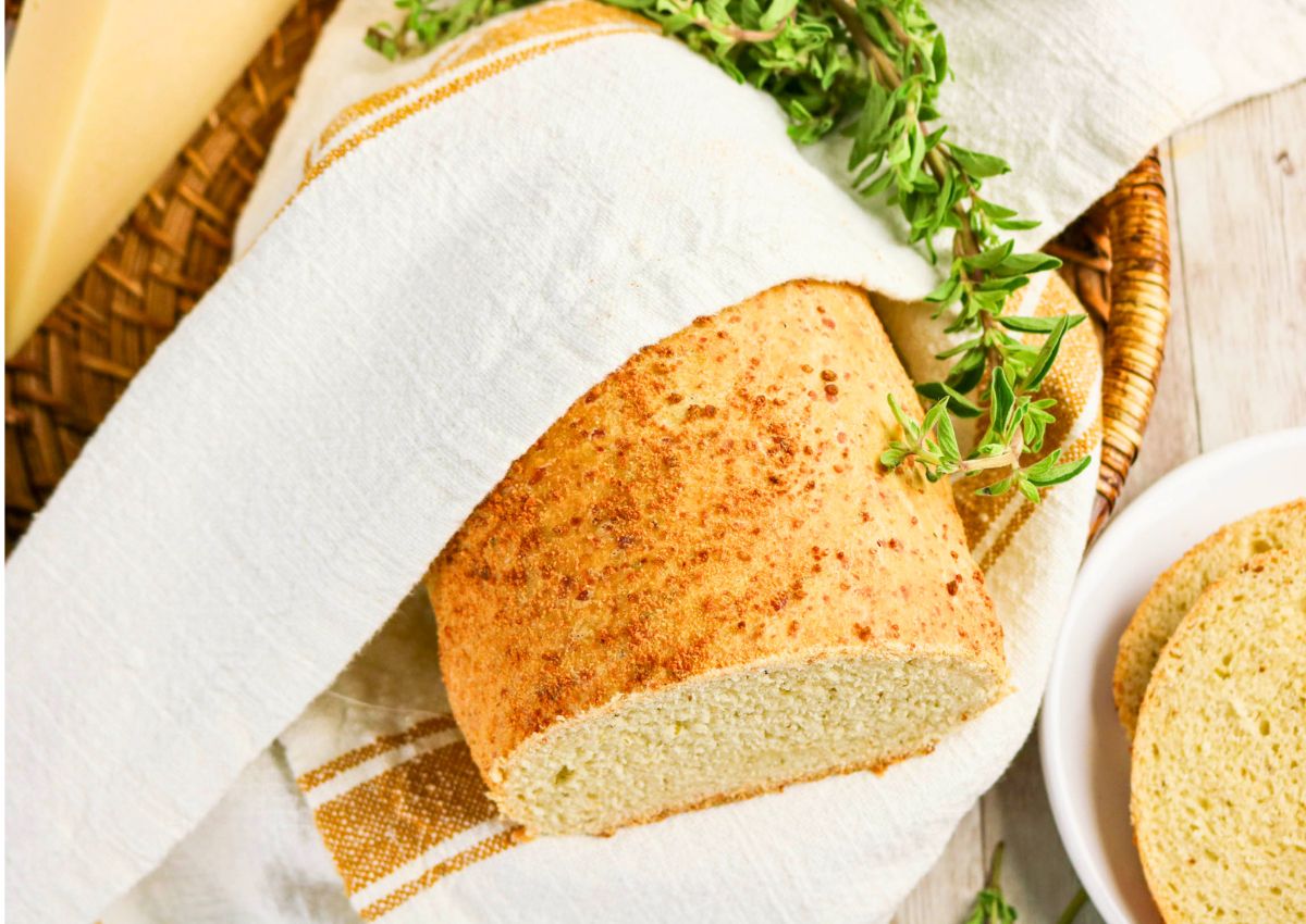 A loaf of bread partially wrapped in a white cloth with a sprig of fresh herbs, placed in a woven basket on a wooden surface.