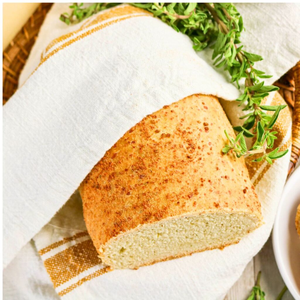 A loaf of bread partially wrapped in a white cloth with a sprig of fresh herbs on top, placed in a woven basket.