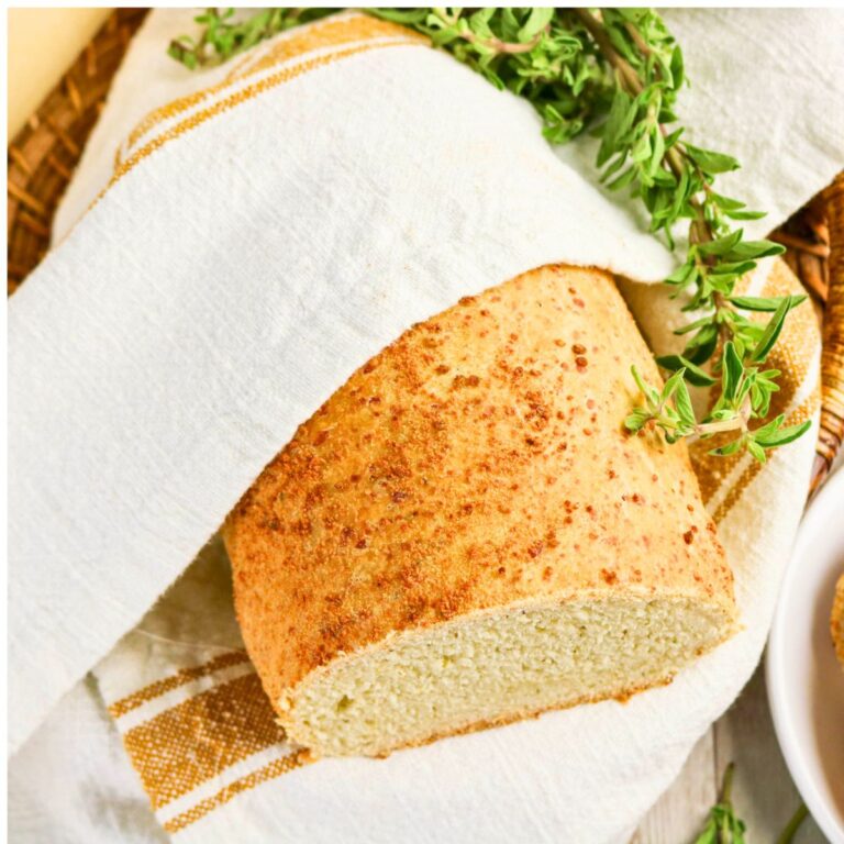 A loaf of bread partially wrapped in a white cloth with a sprig of fresh herbs on top, placed in a woven basket.