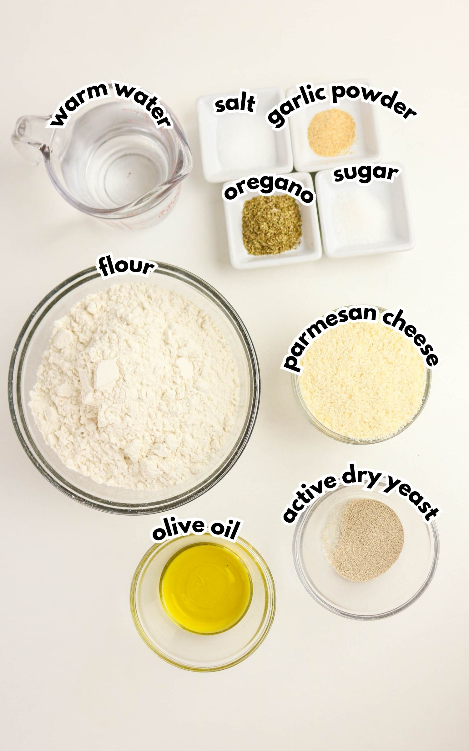 Bowls and dishes containing flour, warm water, olive oil, parmesan cheese, active dry yeast, oregano, garlic powder, salt, and sugar arranged on a white surface.