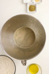 A mixing bowl with yeast and water, surrounded by a bowl of flour, a bowl of olive oil, and two small dishes with dried herbs and seasonings.