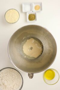 A mixing bowl with yeast and water surrounded by bowls of flour, cornmeal, oil, and small dishes of salt, oregano, and garlic powder on a white surface.