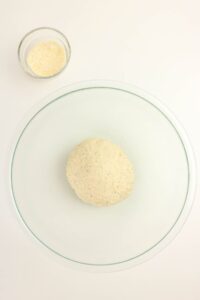 A ball of dough sits in a large glass bowl with a small glass bowl of grated cheese nearby on a white surface.