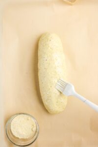 A pastry brush is being used to apply an egg wash to an unbaked loaf of bread dough on parchment paper, with a small bowl of grated cheese nearby.