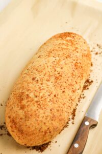A loaf of freshly baked bread with a golden, crisp crust sits on parchment paper next to a bread knife with a wooden handle.