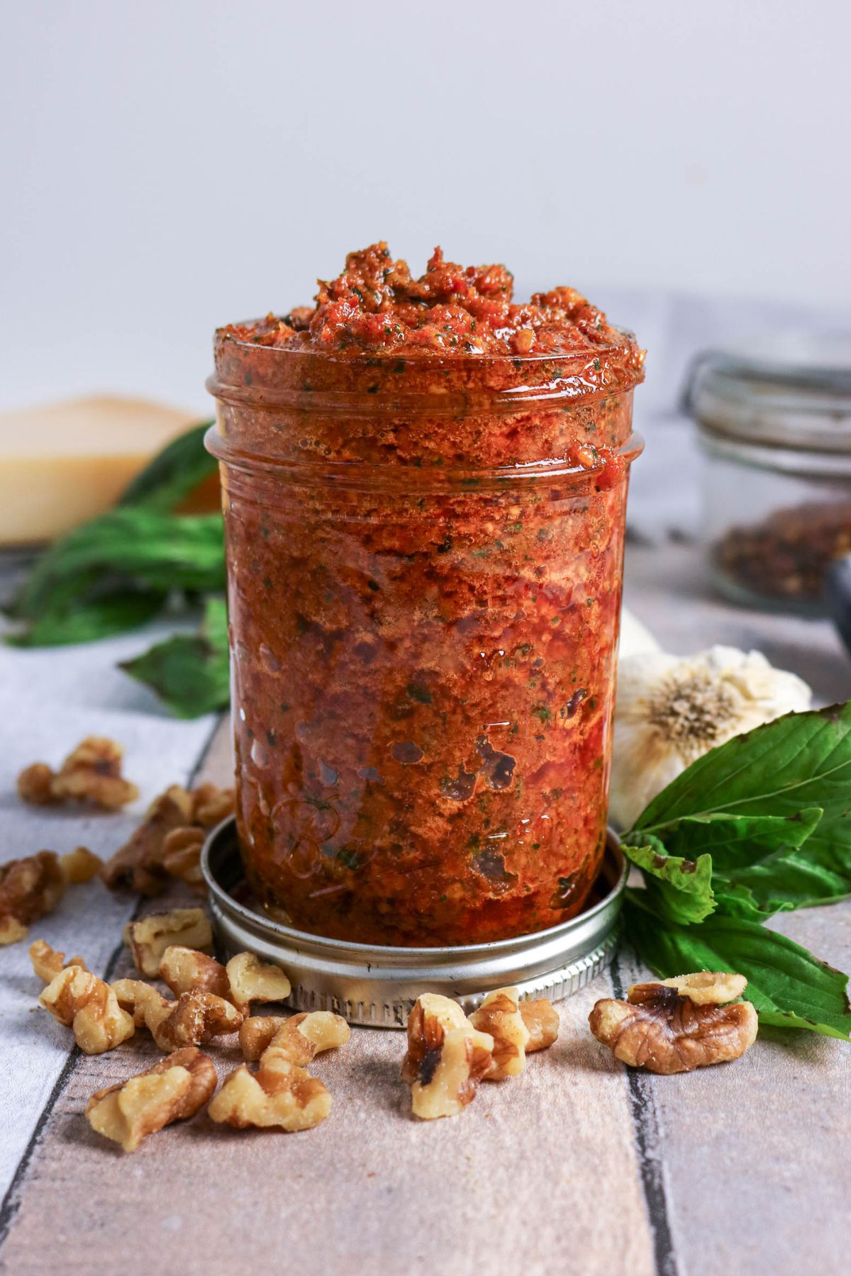 A glass jar filled with chunky red pesto sits on a lid, surrounded by walnuts, basil leaves, and garlic cloves on a wooden surface.