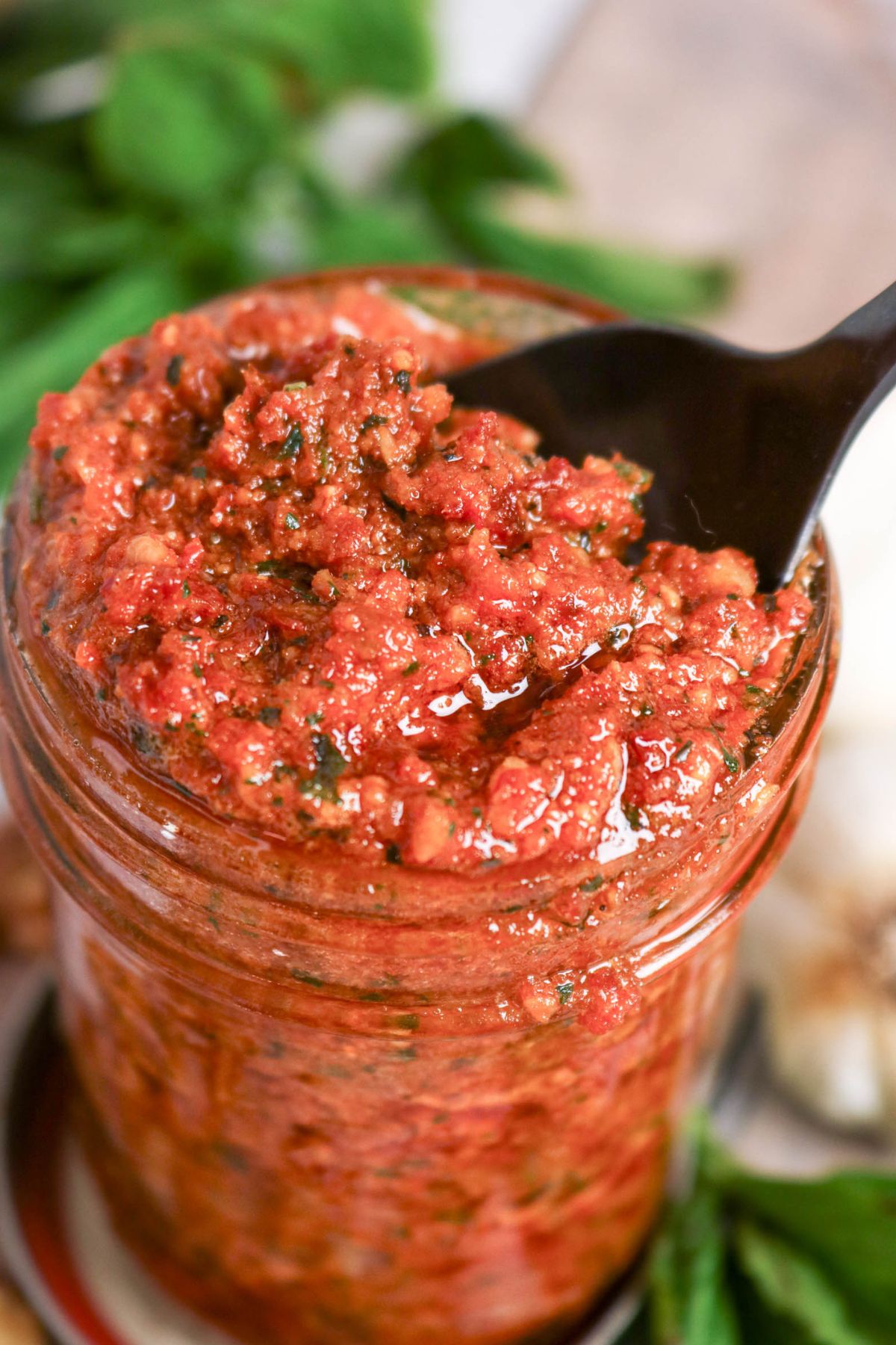 A close-up of a jar filled with chunky red sauce, being scooped with a black spoon. Fresh herbs are visible in the sauce.