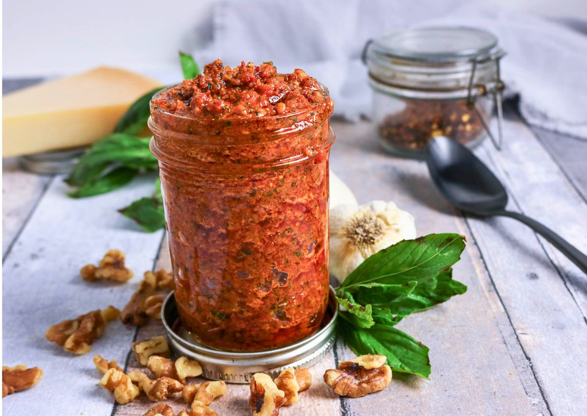 A jar of red pesto sauce on a wooden surface, surrounded by walnuts, basil leaves, garlic, and a spoon, with a closed jar and cheese in the background.