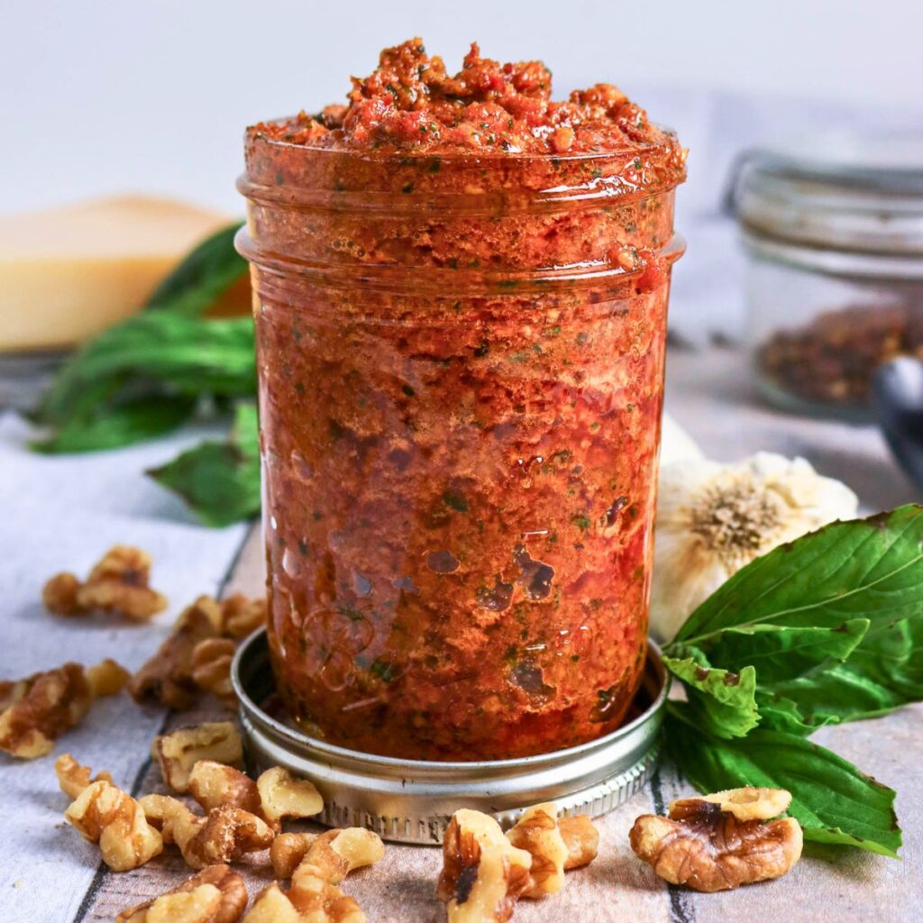 A glass jar filled with red pesto sauce sits on a table, surrounded by walnuts, fresh basil leaves, garlic, and a wedge of cheese in the background.